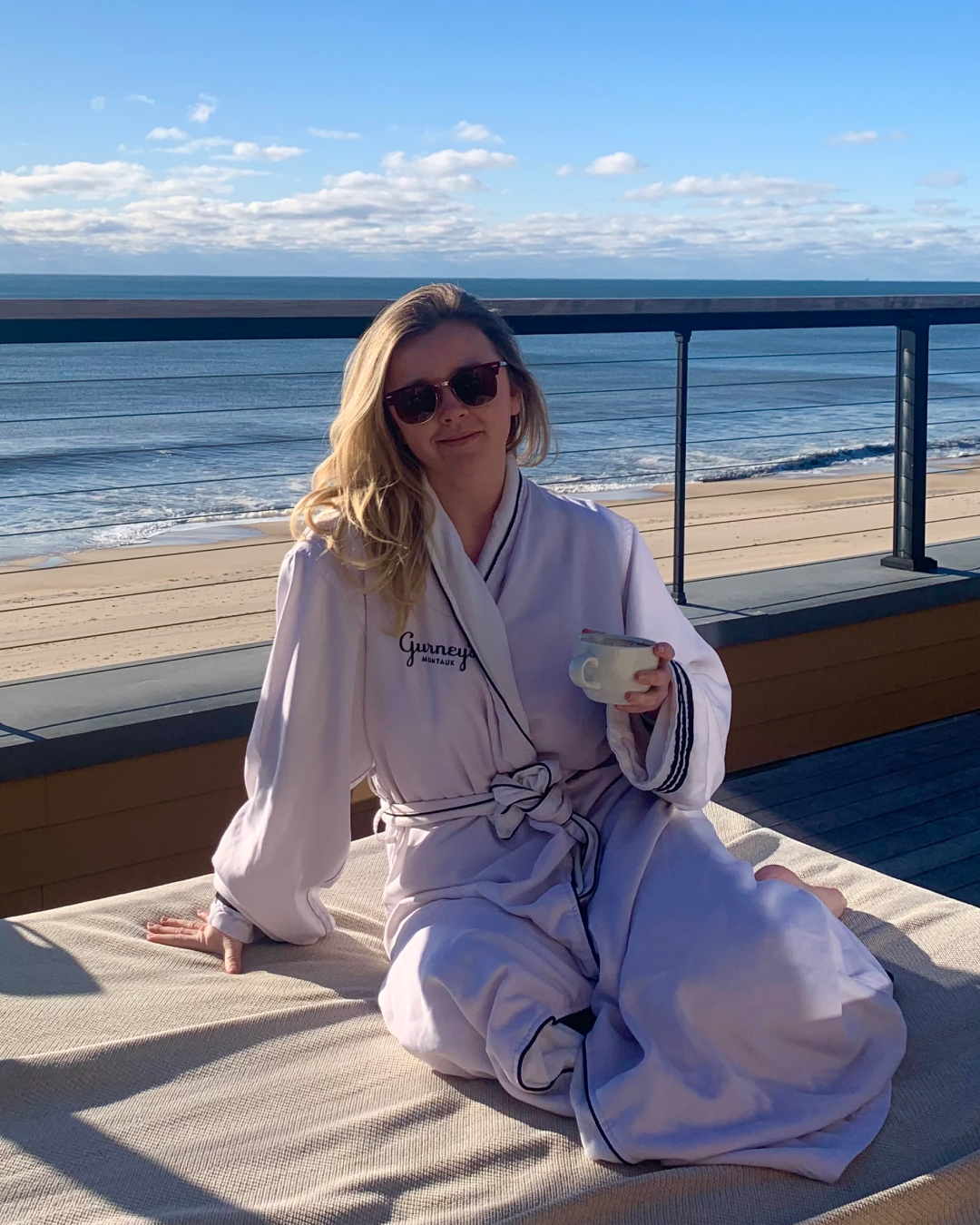 Woman in robe holding a mug, sitting on a deck by the ocean, with a lantern nearby on a sunny day.